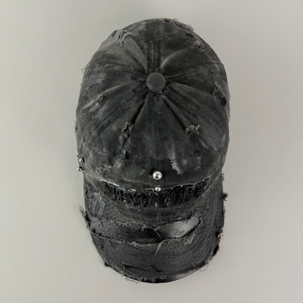 A distressed black baseball cap with frayed edges, distressed detailing, and small silver metalembellishments on the front, photographed against a plain light gray background.