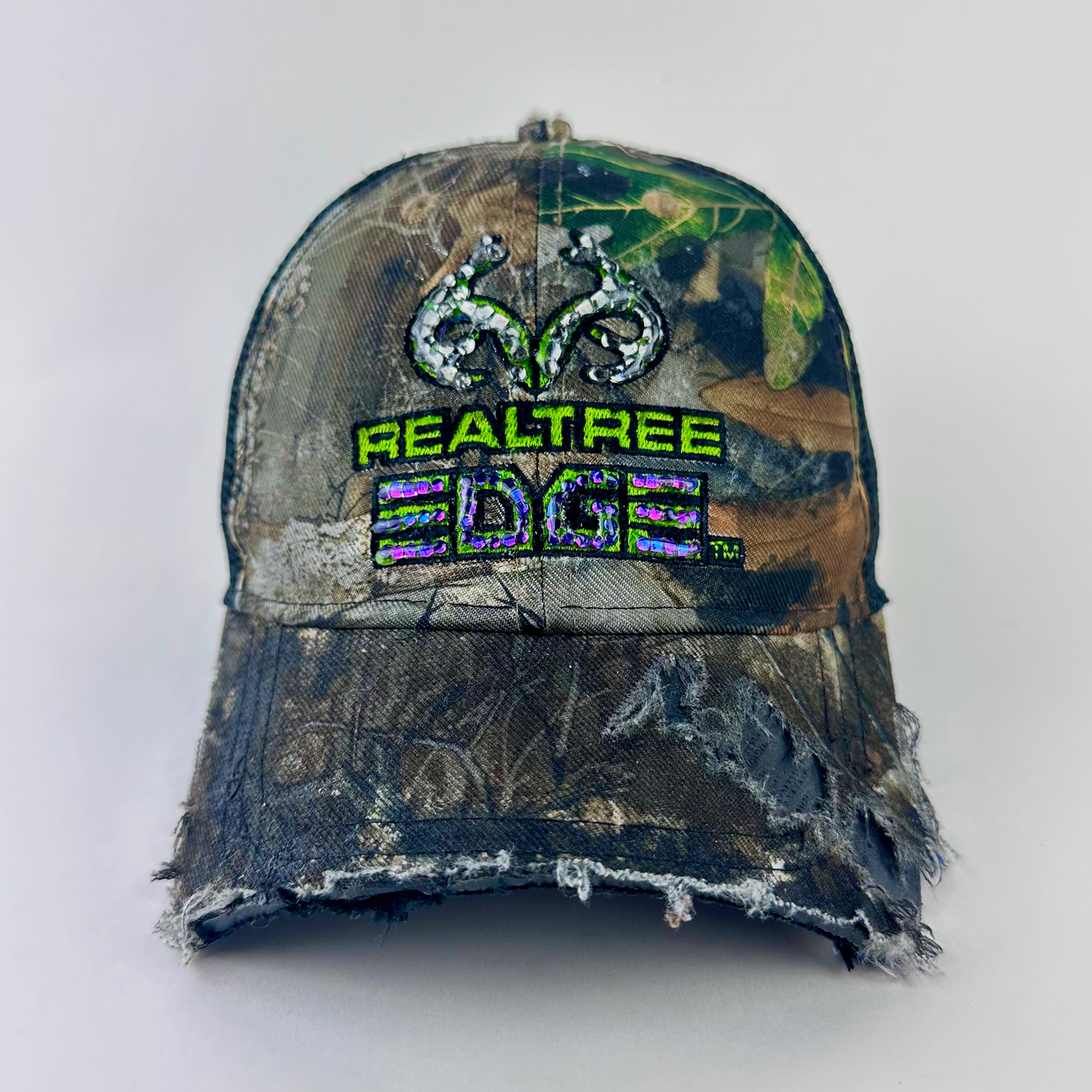 ASTROBOI* x RealTree 'Edge' brown camo rhinestone cap with black and purple/blue rhinestone-embellished RealTree Edge branding across the top and partially sawed-off brim, front view.