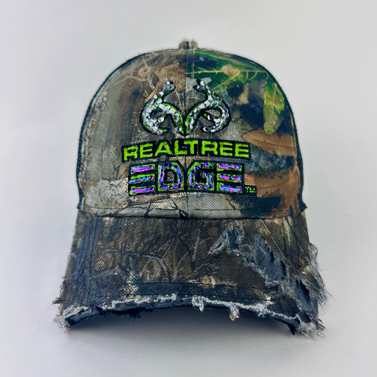 ASTROBOI* x RealTree 'Edge' brown camo rhinestone cap with black and purple/blue rhinestone-embellished RealTree Edge branding across the top and partially sawed-off brim, front view.