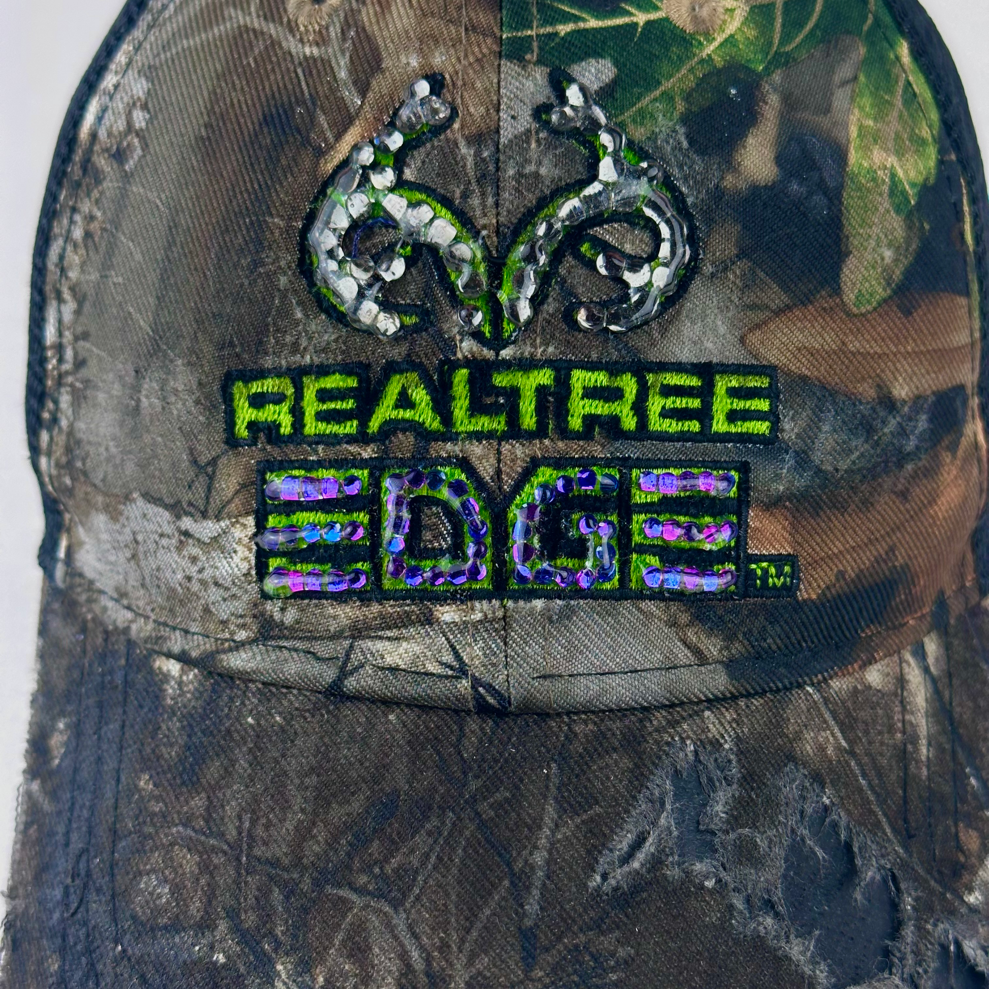 Close-up of ASTROBOI* x RealTree 'Edge' showing black and purple/blue rhinestone embellishment on RealTree Edge branding and custom paint treatment on brown camo fabric.