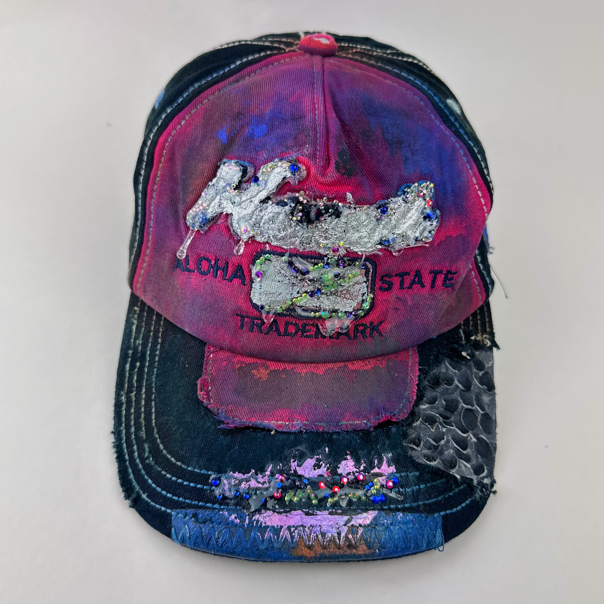 ASTROBOI* 'Tropicale' red and black Y2K Hawaii rhinestone hat with silver embroidered 'Hawaii ALOHA STATE TRADEMARK' text, purple and pink paint splatter, and metallic rhinestone embellishments, front view.