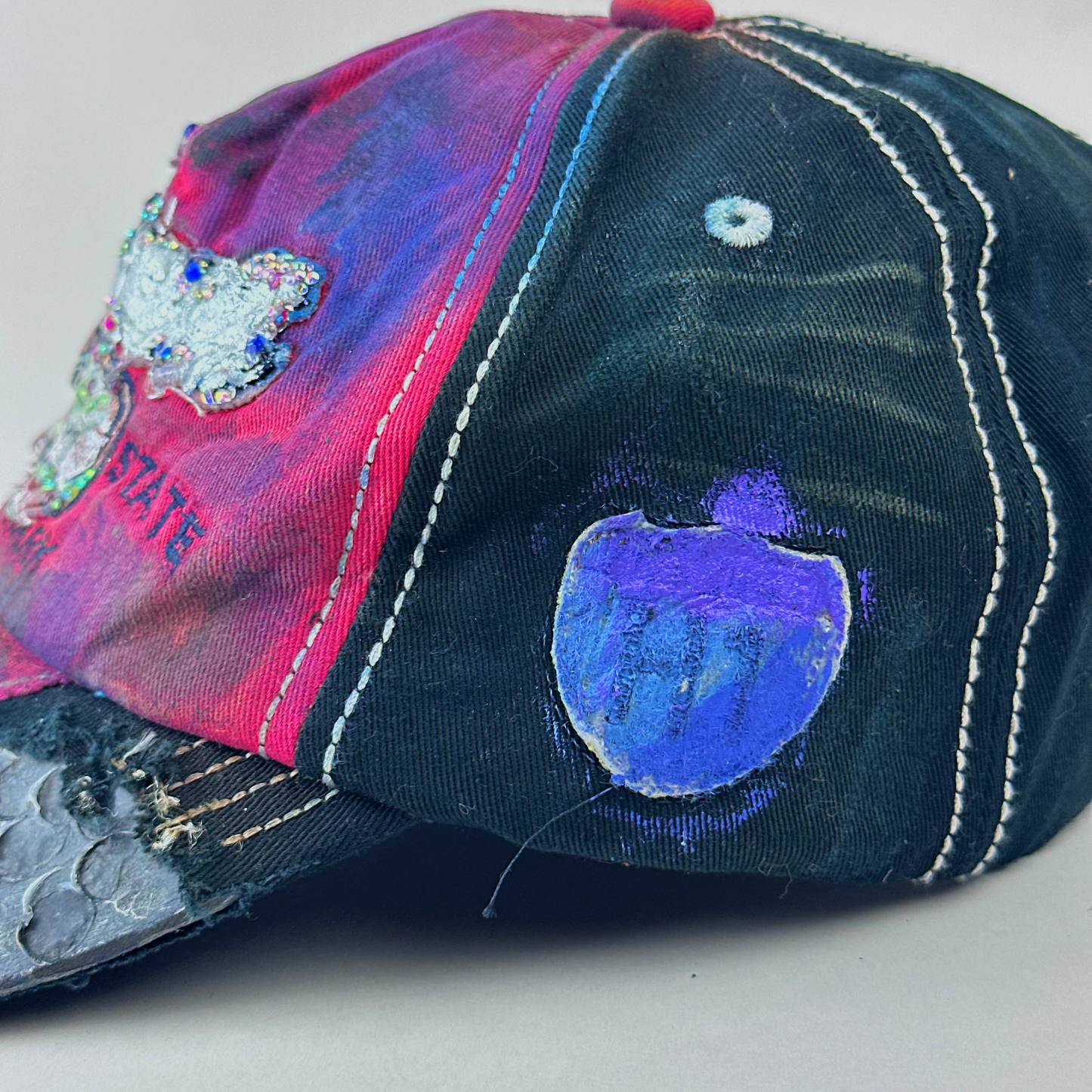 A distressed baseball cap with a split design, featuring a dark washed black denim side with white stitching and a vibrant pink, purple, and blue painted side. The cap has a hand-painted blue shield patch, a glittery silver patch, and a small red button on top.