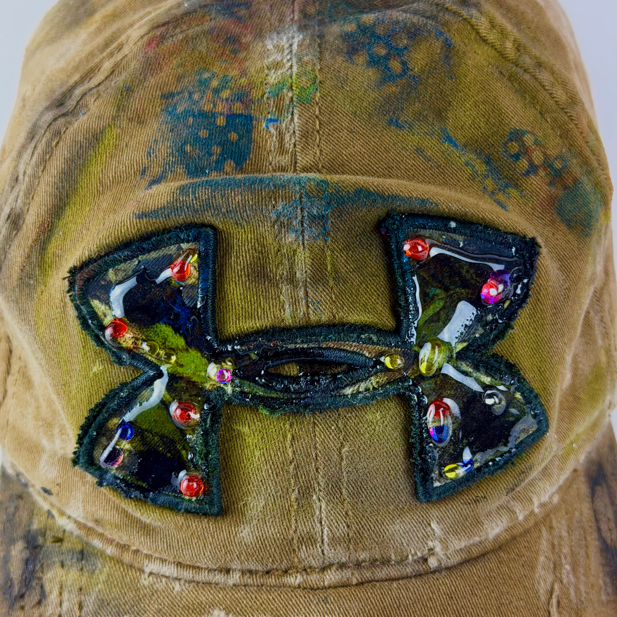 A close-up of a distressed tan baseball cap featuring a large, custom hand-painted Under Armour logo patch on the front. The patch is decorated with colorful rhinestones and metallic embellishments, set against a background of distressed canvas with visible frayed edges.