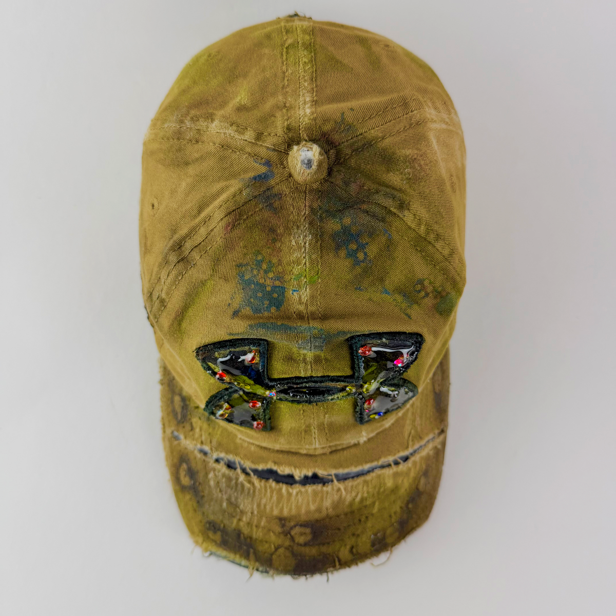 A distressed olive green baseball cap with a heavily embellished Under Armour logo on the front, featuring colorful rhinestones and a frayed, worn finish.