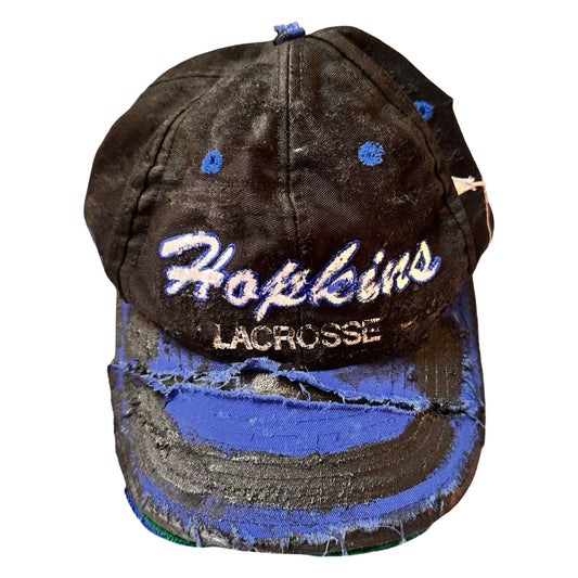 Flat front of Black distressed ASTROBOI* Hopkins Lacrosse cap with blue embroidery and custom black paint treatment, snapback style, handcrafted in NYC.