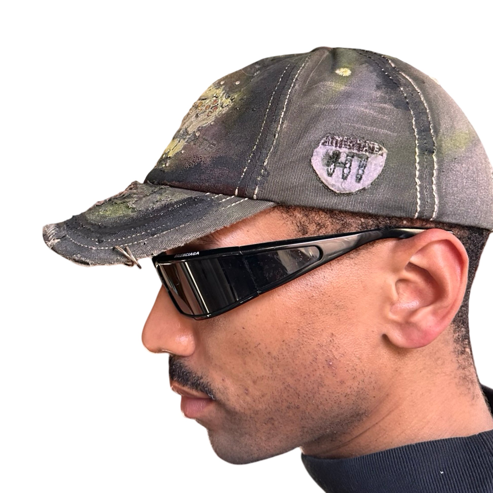 Left of “Green and brown distressed ASTROBOI* Hawaii hat with rhinestones, metallic paint, and sterling-steel piercings, customized in NYC.”