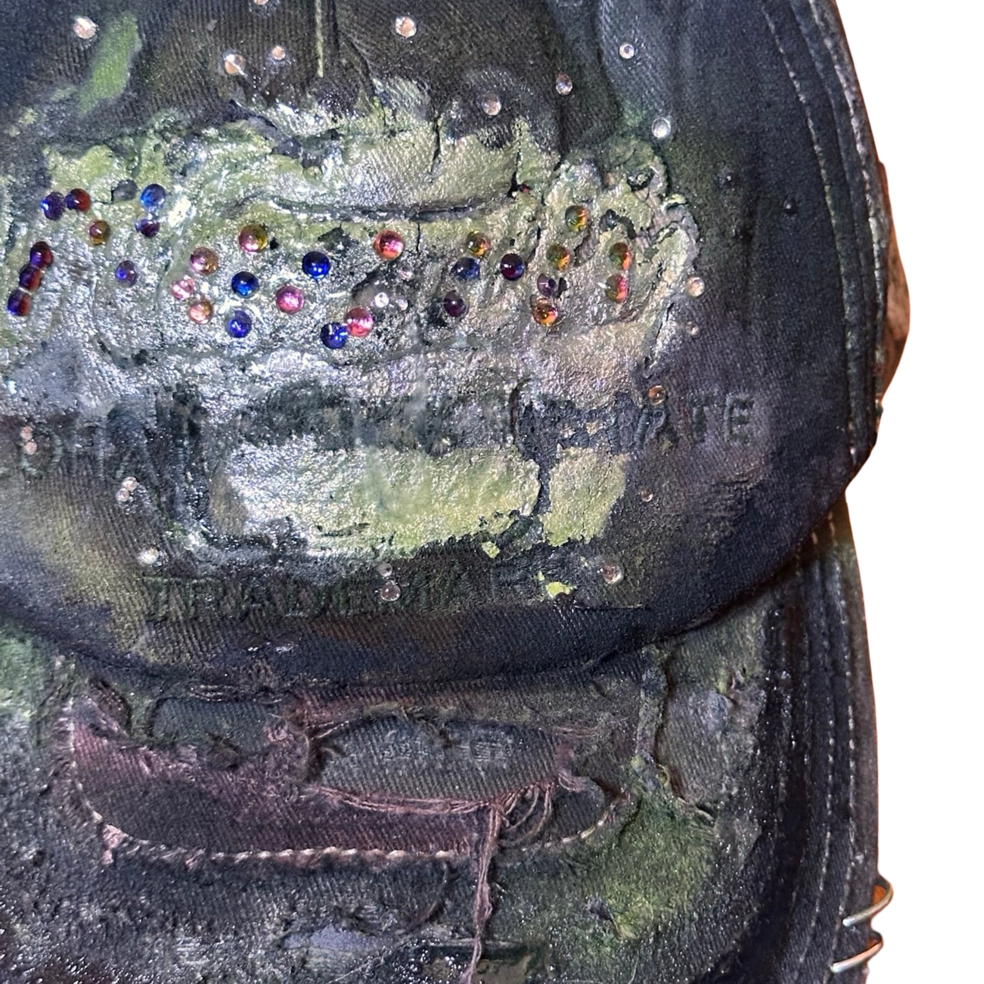 Flat close up image of “Green and brown distressed ASTROBOI* Hawaii hat with rhinestones, metallic paint, and sterling-steel piercings, customized in NYC.”
